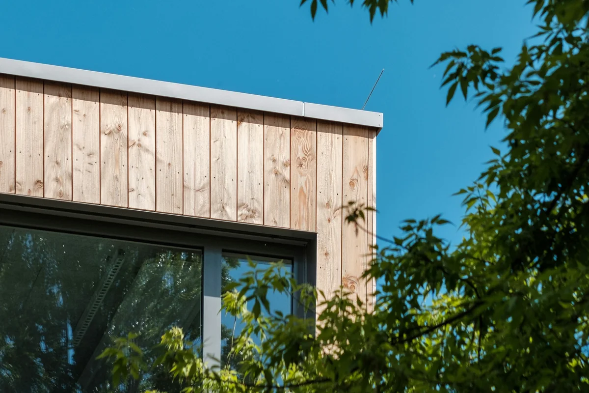 Angle d'une façade en bardage bois vertical clair sous un ciel bleu, avec une baie vitrée et un feuillage au premier plan