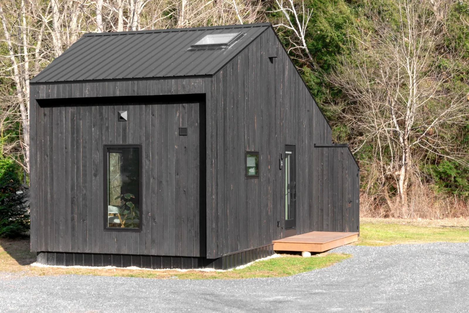 Petite maison contemporaine à bardage bois vertical teinté noir et toit en métal, avec une marche d'entrée en bois, au bord d'une allée gravillonnée