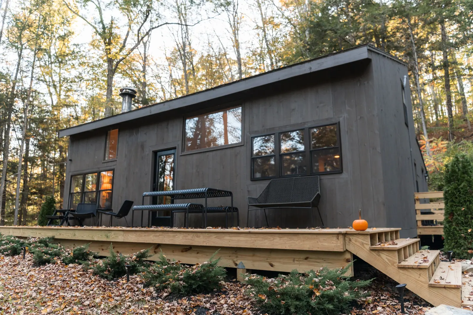 Maison bois de plain-pied, longue et sombre, avec une terrasse en bois meublée et un escalier, au milieu d'arbres en automne