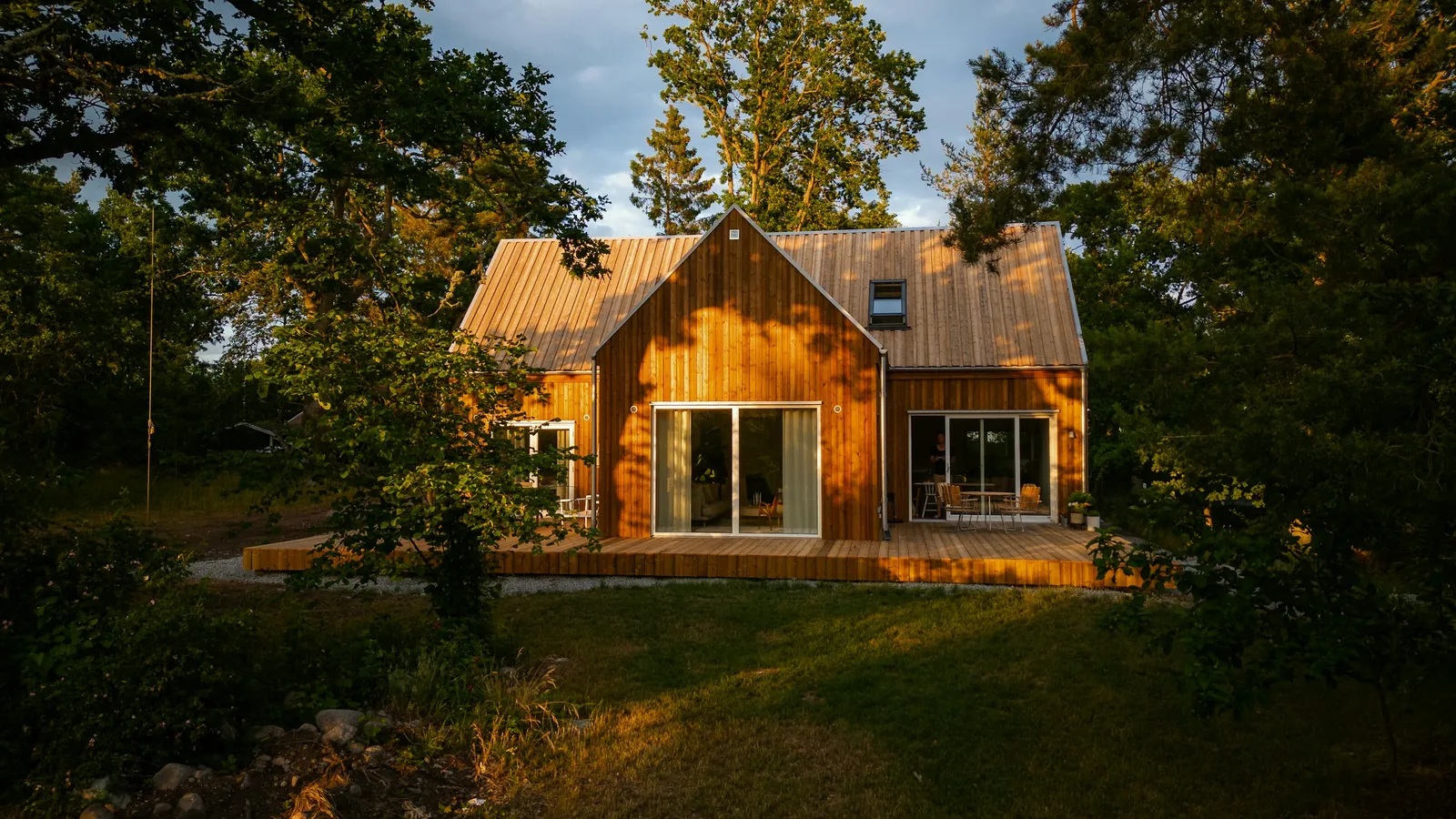 Maison à bardage bois vertical et toit à deux pans, avec une grande terrasse en bois, au milieu d'un jardin arboré dans la lumière du soir