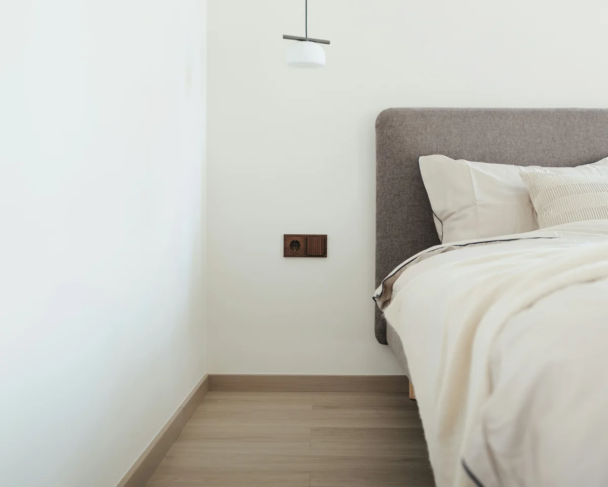 Chambre sobre aux murs blancs, avec un lit à tête de lit grise, une suspension blanche et un sol clair