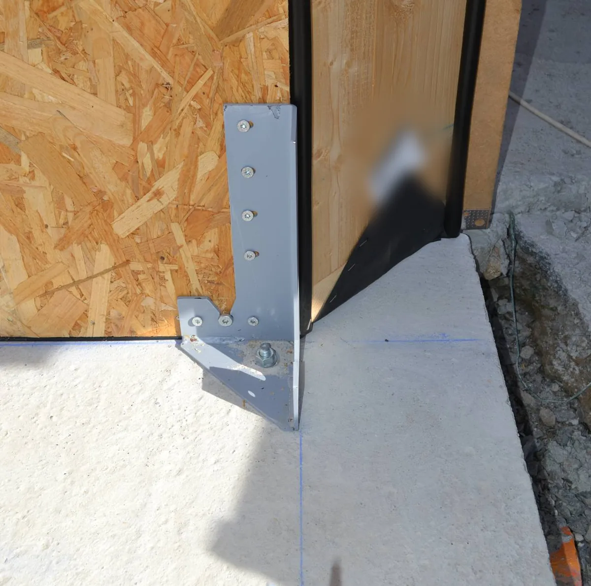 Gros plan d'une équerre métallique vissée au pied d'un mur à ossature bois et boulonnée dans la dalle béton