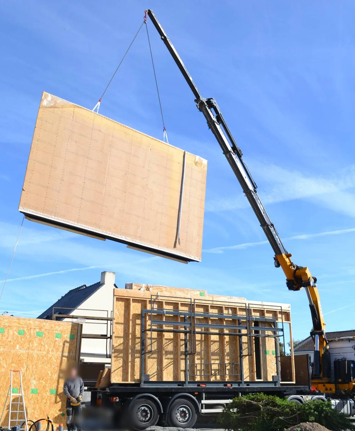 Une grue de camion soulève un grand panneau mural à ossature bois au-dessus d'une remorque chargée d'autres panneaux, à côté d'une dalle béton