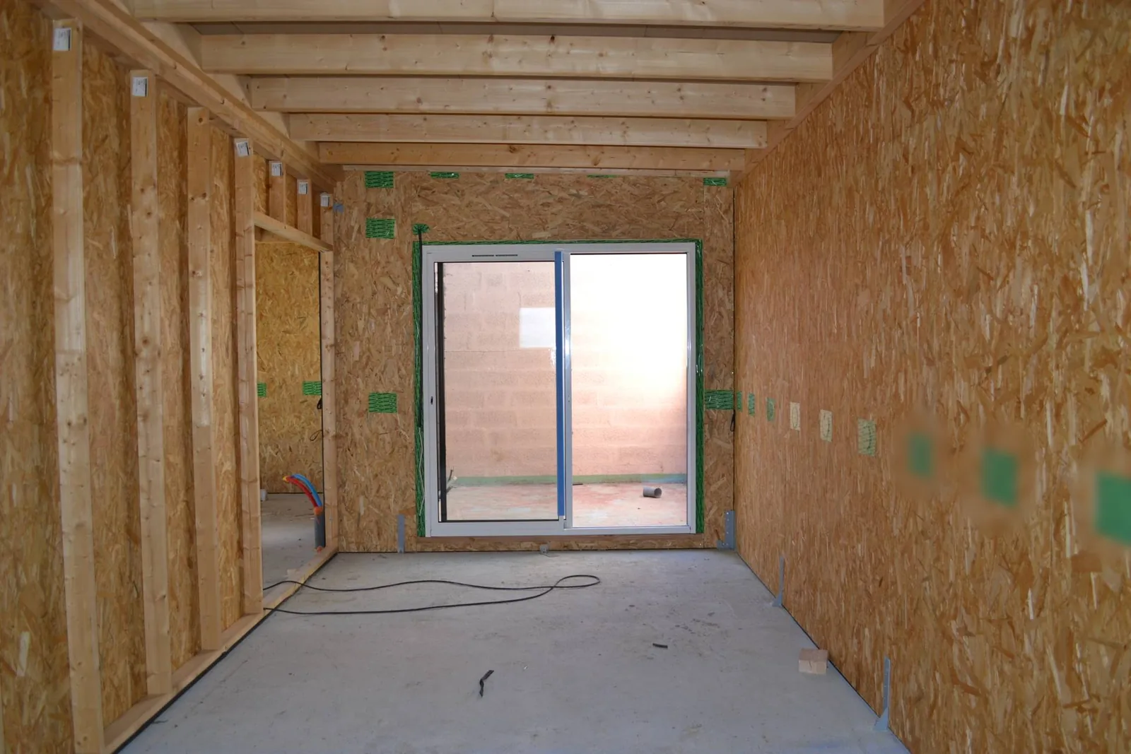 Pièce en chantier vue de l'intérieur : murs en panneaux OSB, solives en bois au plafond, sol en dalle béton brute et baie vitrée déjà posée